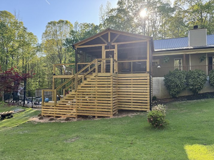 A wooden deck with stairs and a screened-in porch attached to a house, surrounded by green grass and trees, during daytime.