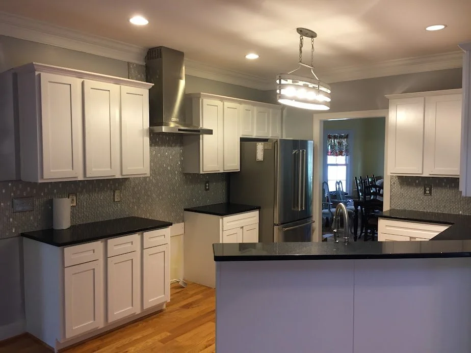 Kitchen with white cabinets, black countertops, stainless steel refrigerator, and mosaic tile backsplash, with a view into a dining area with a dark wooden table and chairs.