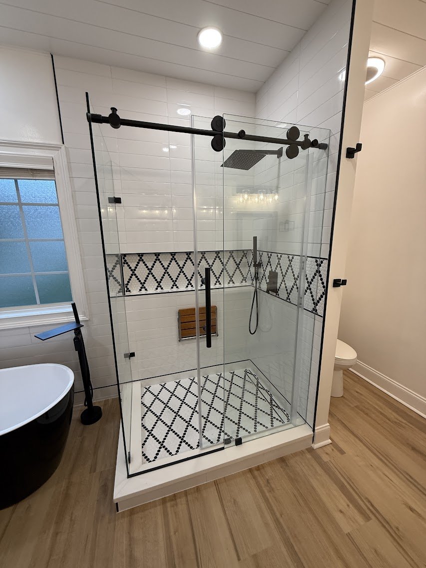 A modern enclosed glass shower with black hardware and a built-in bench, next to a white toilet and a black bathtub, with a window on the left and wood flooring.