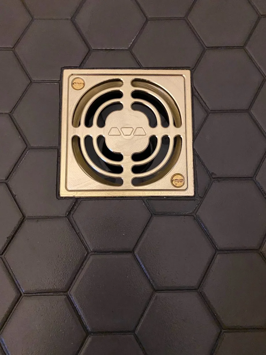 Floor drain with a metal grate cover on hexagonal black tile flooring.