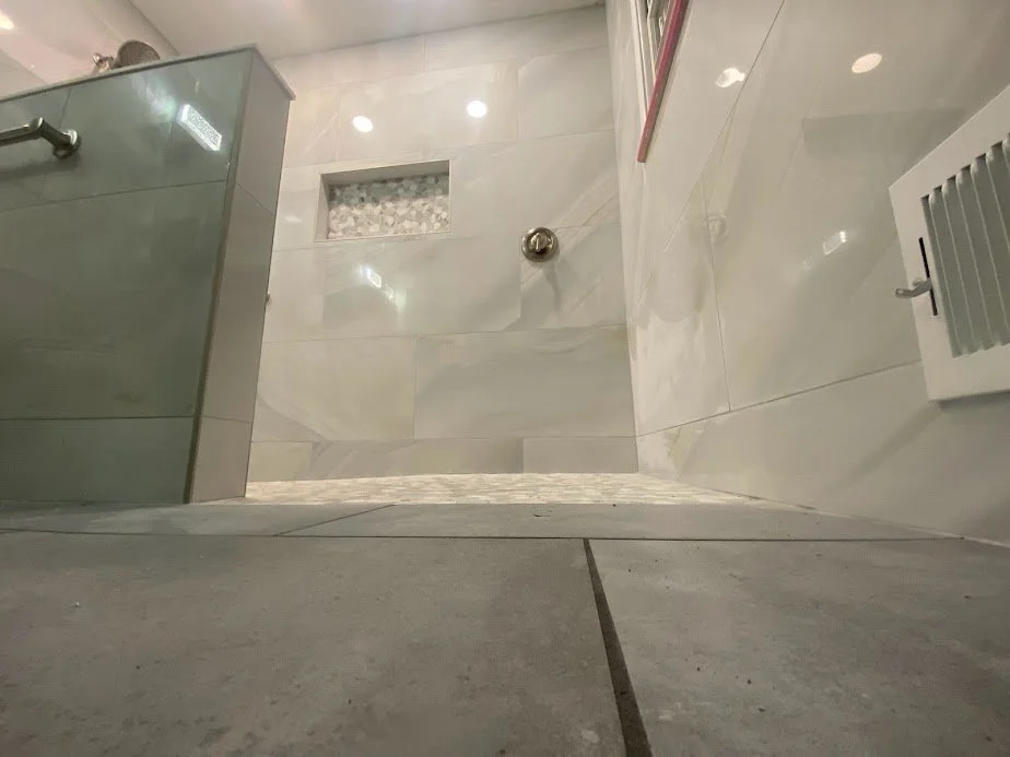 View of a bathroom shower with white marble tiles, a glass shower door, a wall-mounted showerhead, a small recessed shelf with decorative stones, and bathroom fixtures on the wall.