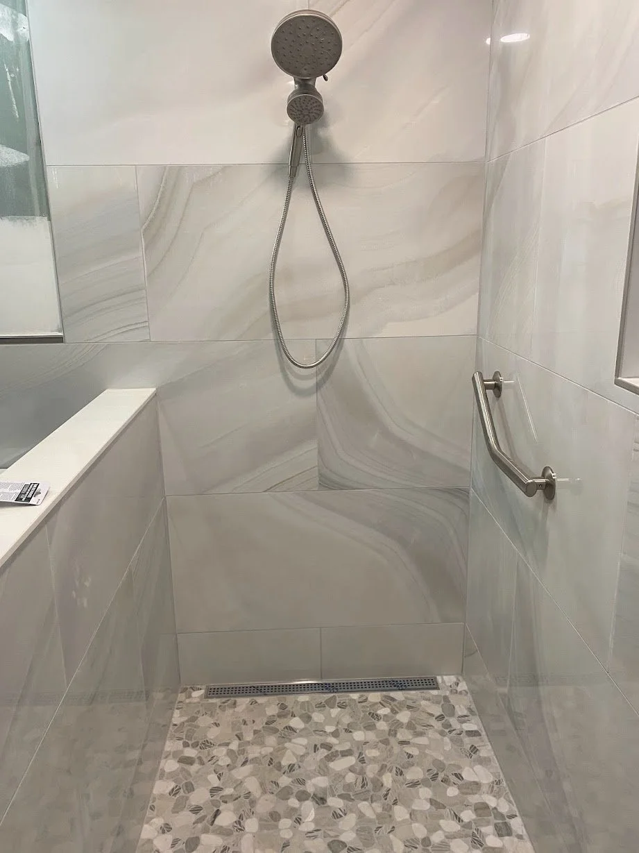 Shower area with marble-patterned tiles, a handheld showerhead, a grab bar, and a tiled floor with pebble design.