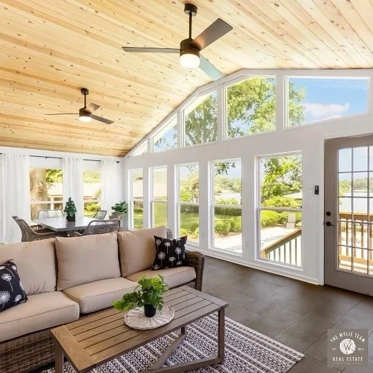 Bright sunroom with large windows showing green trees and outdoor patio, beige sectional sofa with black and white throw pillows, wooden coffee table with a small plant, ceiling fans, and a sliding door leading outside.