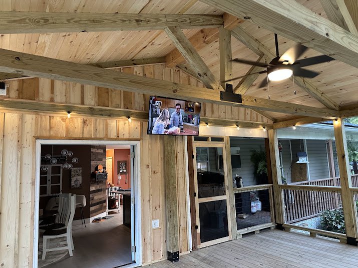 Wooden porch or deck area with a ceiling fan and a mounted TV showing two people talking, adjacent to a room with a dining area and a kitchen.