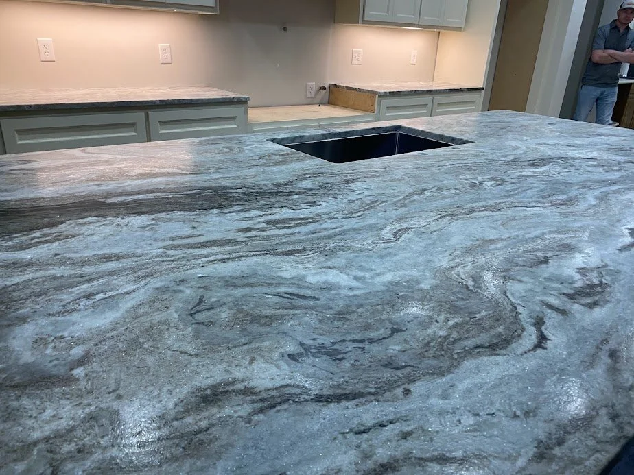 Close-up of a kitchen countertop made of gray marble with white and black veining, and a black undermount sink. In the background, light-colored cabinets and a person standing near the entrance.