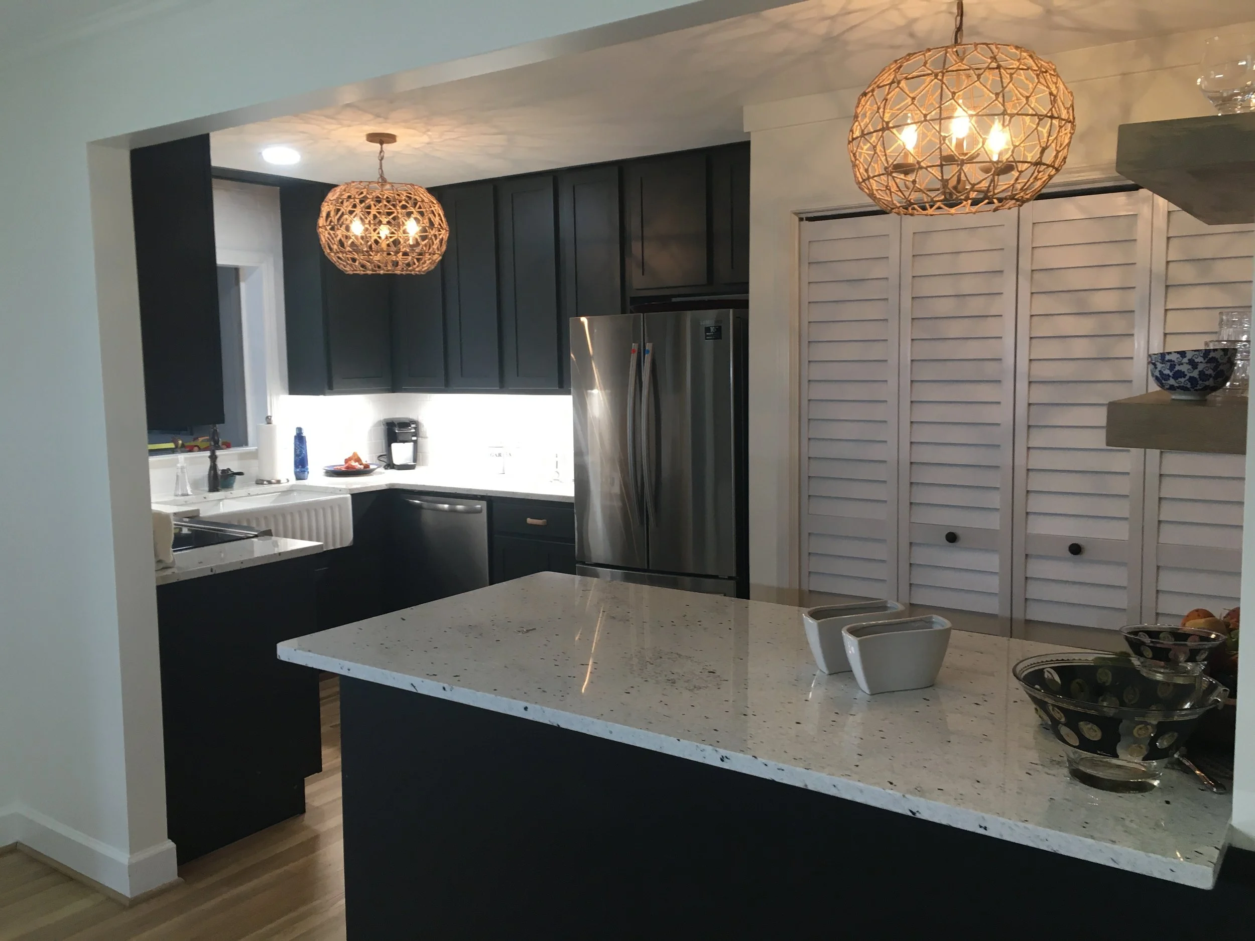 A modern kitchen with white and black cabinets, a white speckled countertop island, stainless steel appliances including a double-door refrigerator, two pendant lights with woven shades, a window with a white frame and sink underneath, and a louvered white closet door.