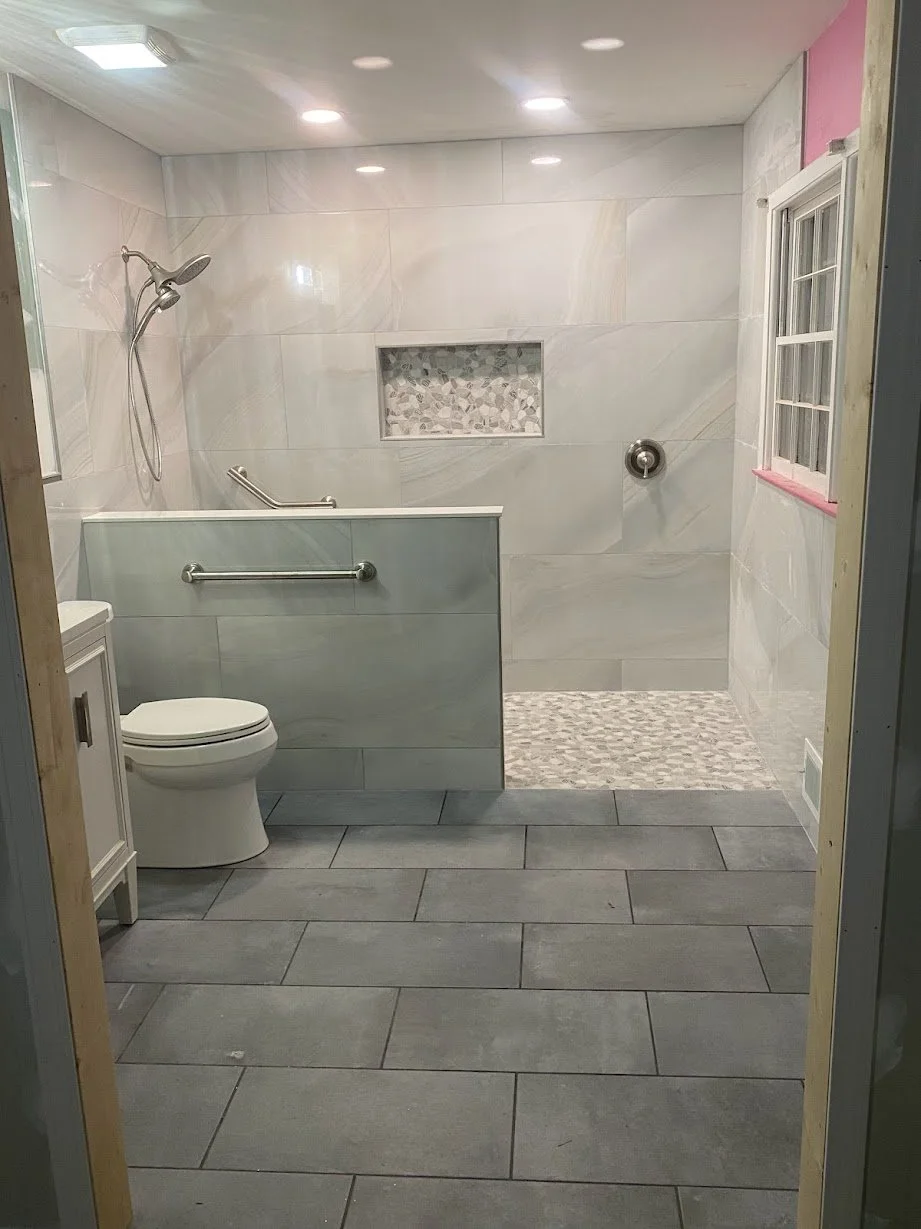A bathroom with a walk-in shower, a toilet, and a small vanity. The shower has a handheld showerhead, a built-in niche with decorative tiles, and a partial glass divider. The floor has large gray tiles, and the shower area has smaller pebble-style ti