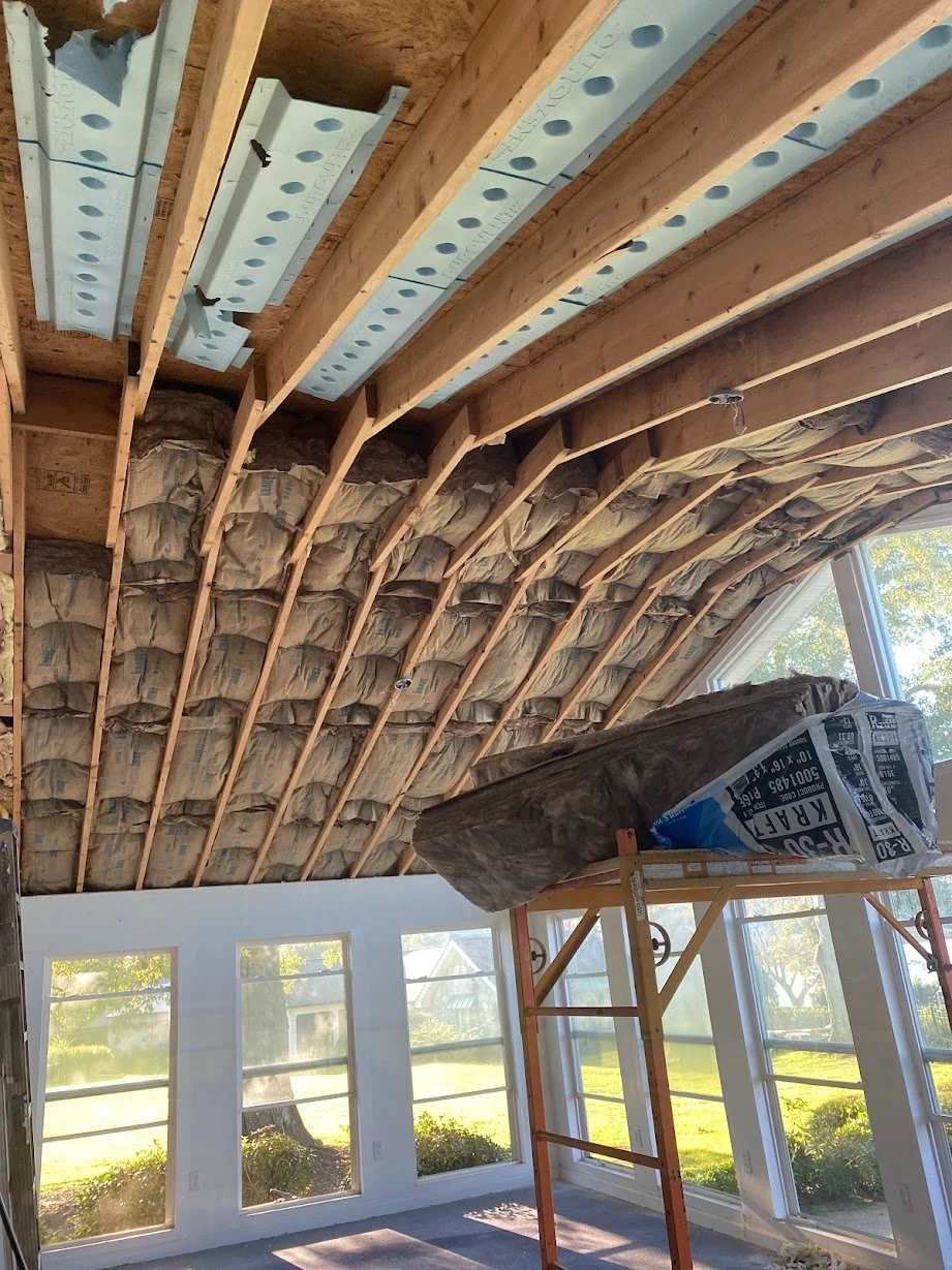 Interior room under construction with insulation being installed in the sloped ceiling, scaffolding, and sunlight through large windows.