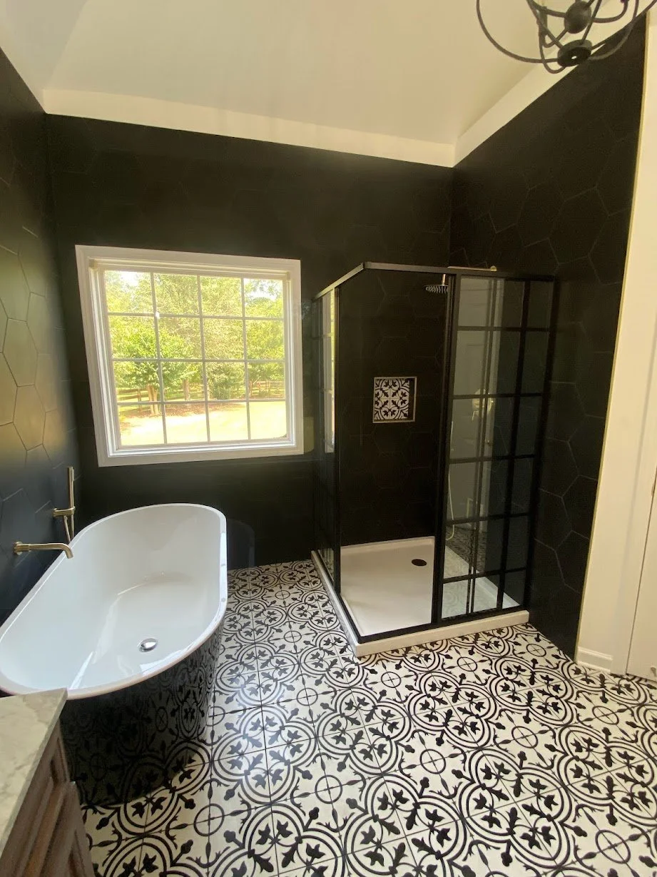 Bathroom with black walls and patterned black and white tile floor, white freestanding bathtub with gold fixtures, black-framed glass shower enclosure, and a window showing green trees outside.