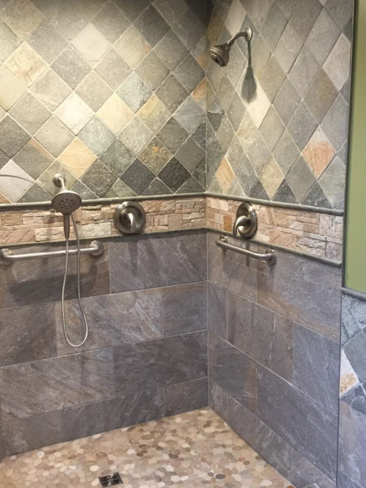 A shower area with multi-colored tile walls, grab bars, a showerhead, and a pebble floor.