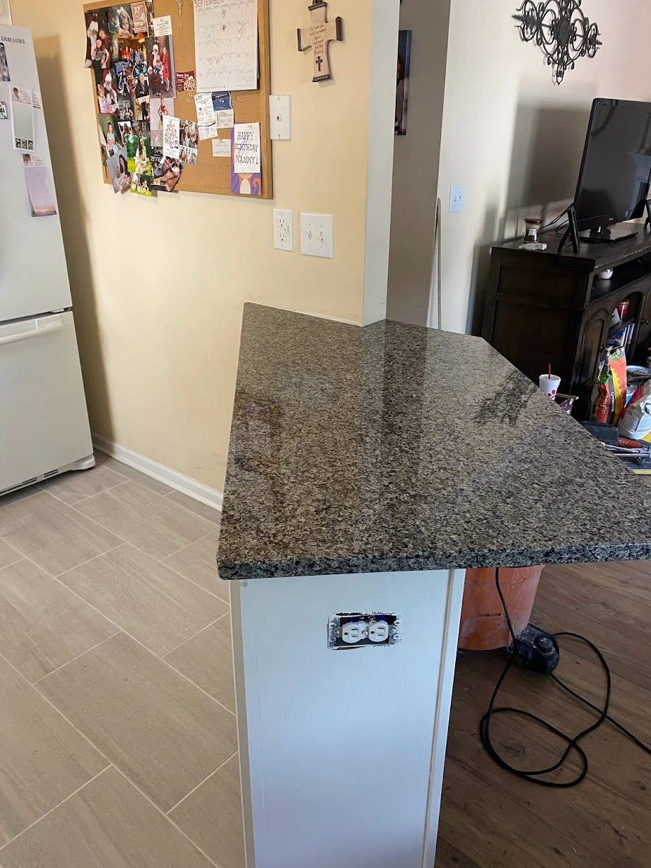 Close-up of a kitchen countertop with electrical outlets on the side of the island, and a living room with a TV and dark cabinet in the background.