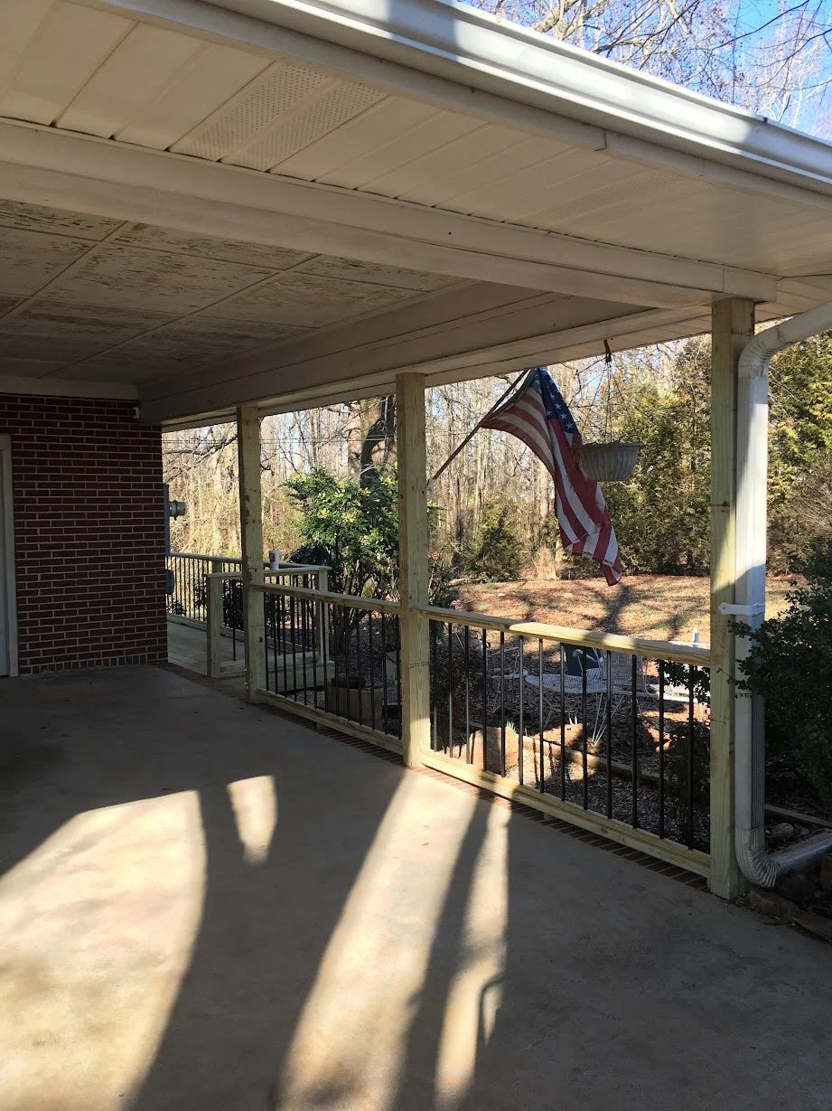 View of a screened porch with a concrete floor, white ceiling, and an American flag hanging from a post. The porch overlooks a backyard with trees and bushes.
