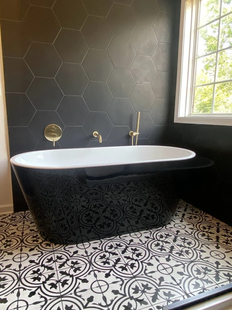 Bathroom with a black clawfoot bathtub, black wall tiles with a hexagon pattern, a large window with sunlight, and patterned black and white floor tiles.