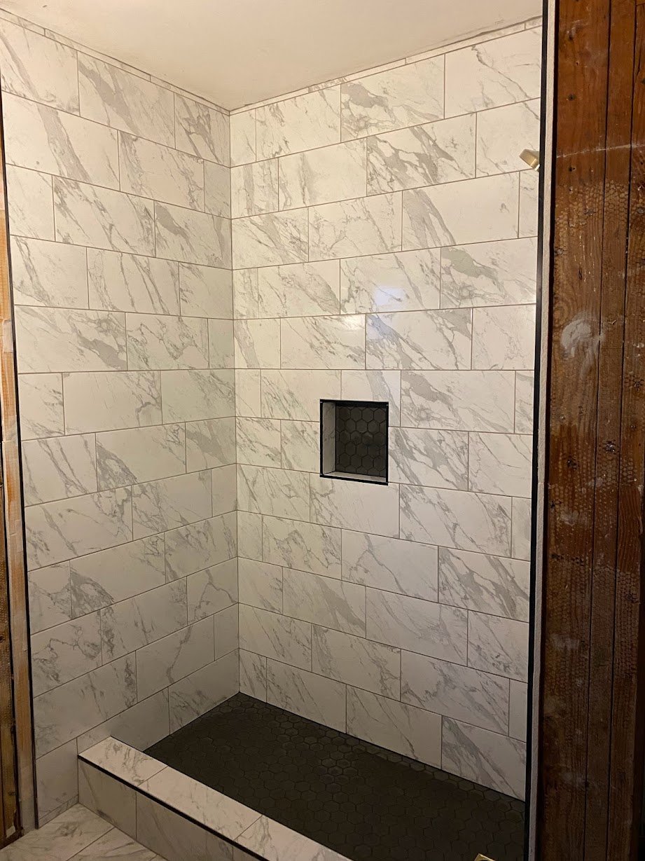 A modern walk-in shower with white marble tiles on the walls, a built-in niche with dark tiles, and small dark hexagonal tiles on the shower floor, bordered by a marble threshold. To the right, the wall is partially wood-paneled.