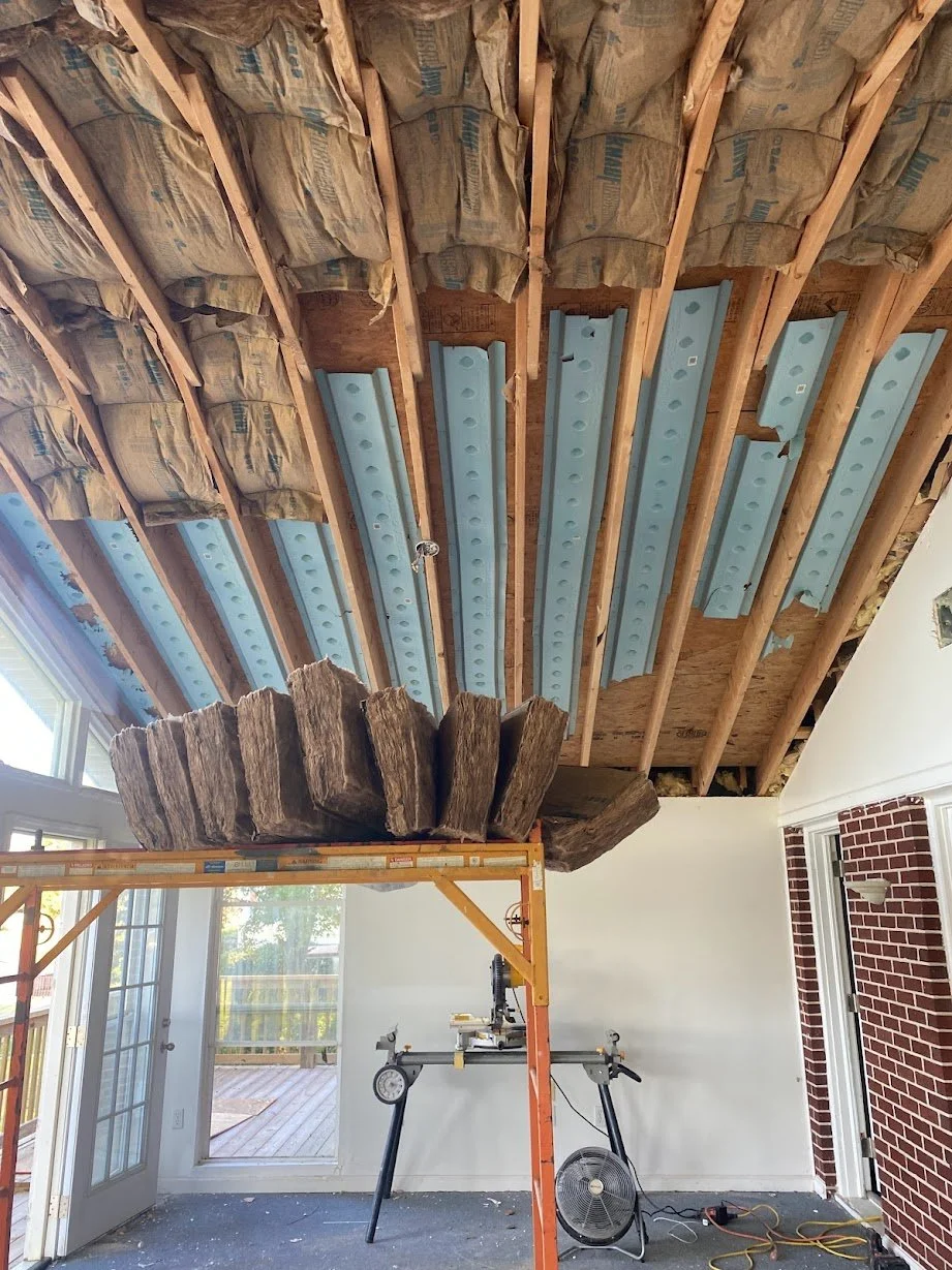 Interior of a house under renovation with exposed ceiling beams, insulation, and metal framing. A large wooden beam is being lifted or supported, and construction tools are present.