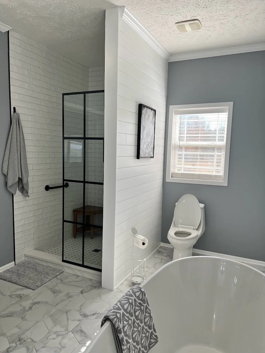 Bathroom with a corner shower featuring black metal framing, white subway tile walls, and a small wooden bench inside. Adjacent to the shower is a toilet near a window with white blinds. A bathtub with a gray patterned towel draped over its edge is p