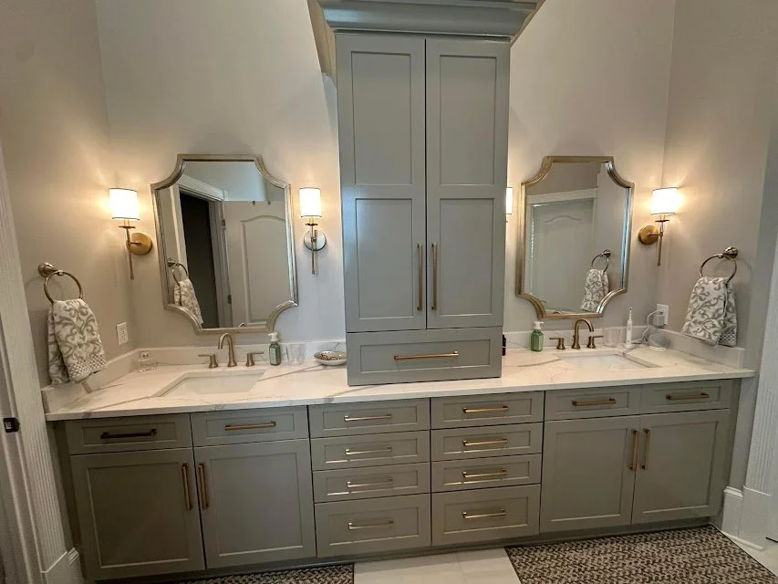 A double bathroom vanity with two mirrors, two sinks, and gold fixtures. Centered above with a tall cabinet and light fixtures on each side of the mirrors.
