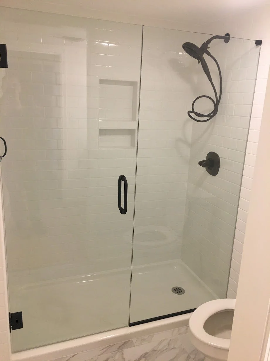 A bathroom shower with a glass door, white tiled walls, a black showerhead, and a toilet in front of the shower.