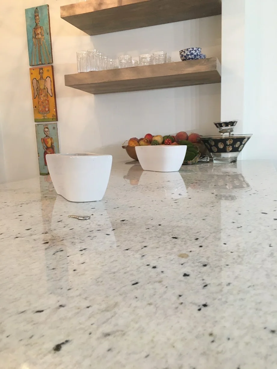 Close-up view of a kitchen counter with white bowls, a bowl of assorted fruits, and decorative bowls, with wooden shelves and artwork on the wall behind.