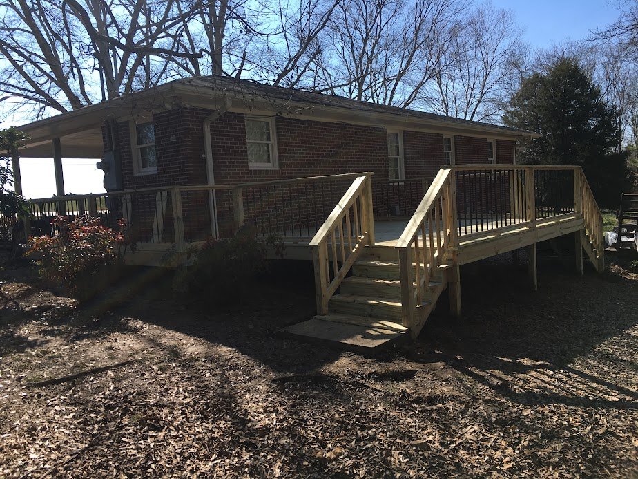 A brick house with a newly built wooden wheelchair-accessible ramp in the backyard. The ramp has black metal railings and stairs at the bottom. Bare trees are visible in the background, and the ground is covered with fallen leaves.