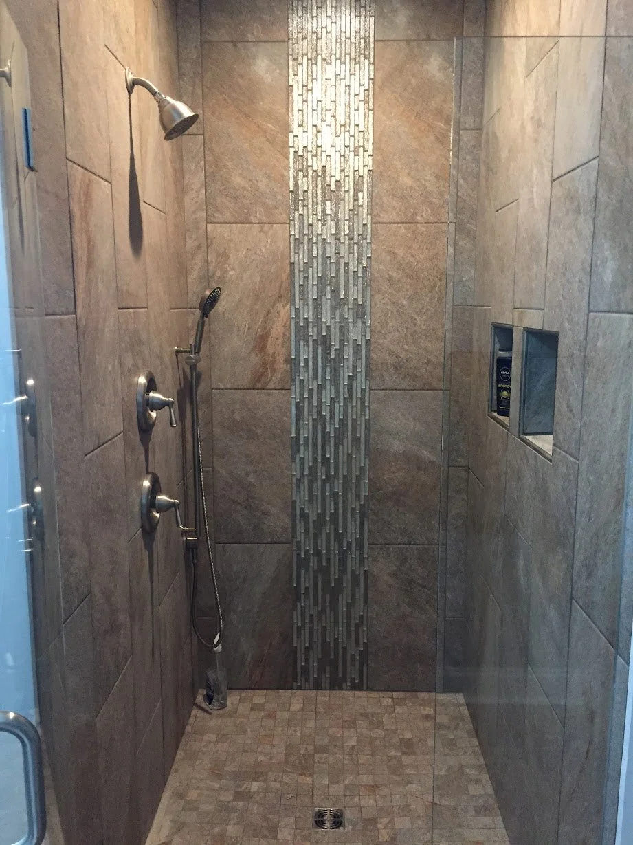 Empty walk-in shower with tan tile walls, a decorative vertical tile strip, and dual shower controls including a handheld showerhead, spray, and a fixed showerhead. Shower niche on the right wall with personal care products, and a floor drain at the 