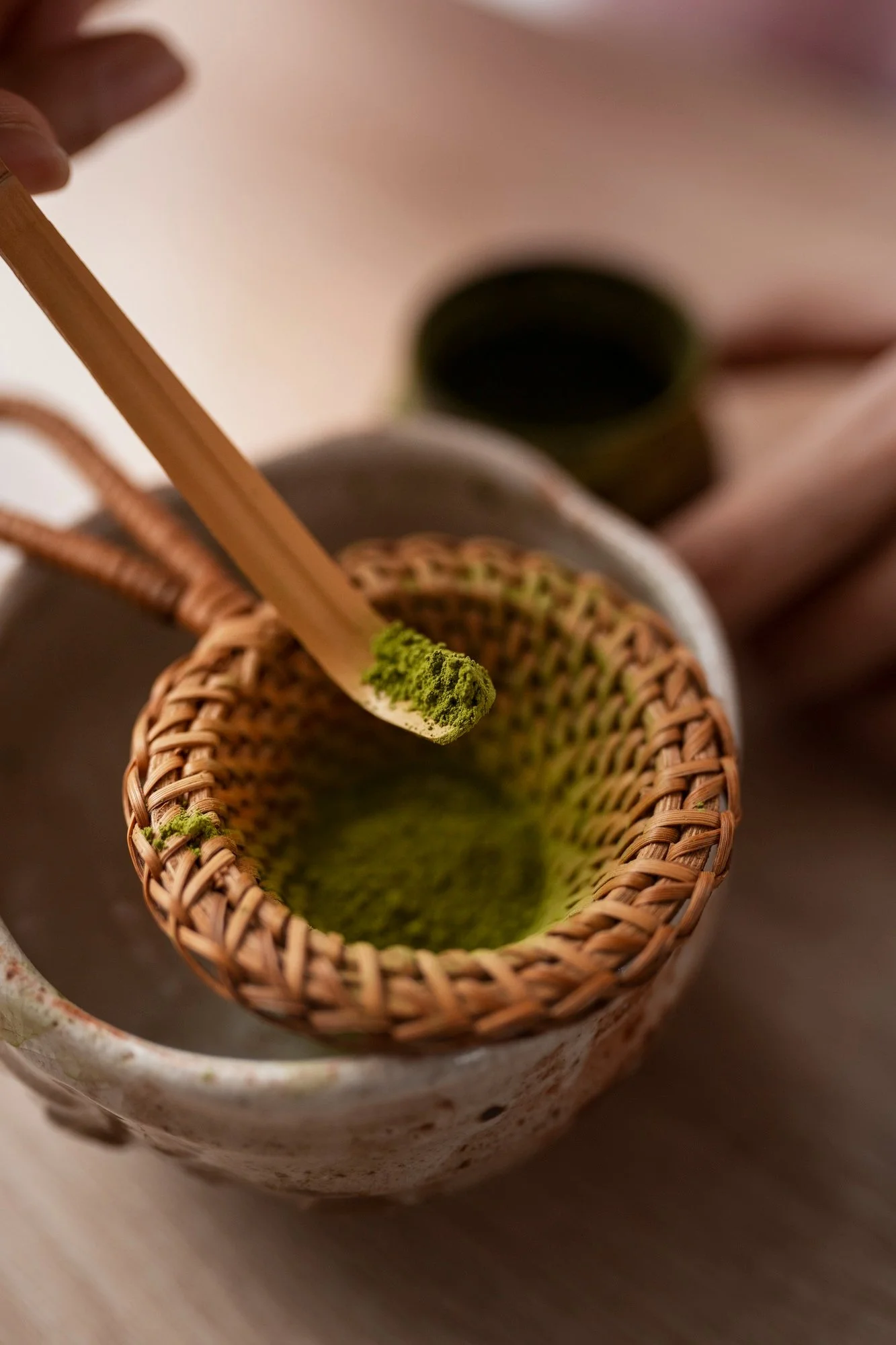 A hand holding a bamboo spatula with green matcha powder over a woven basket, a ceramic cup, and a small bowl in the background.