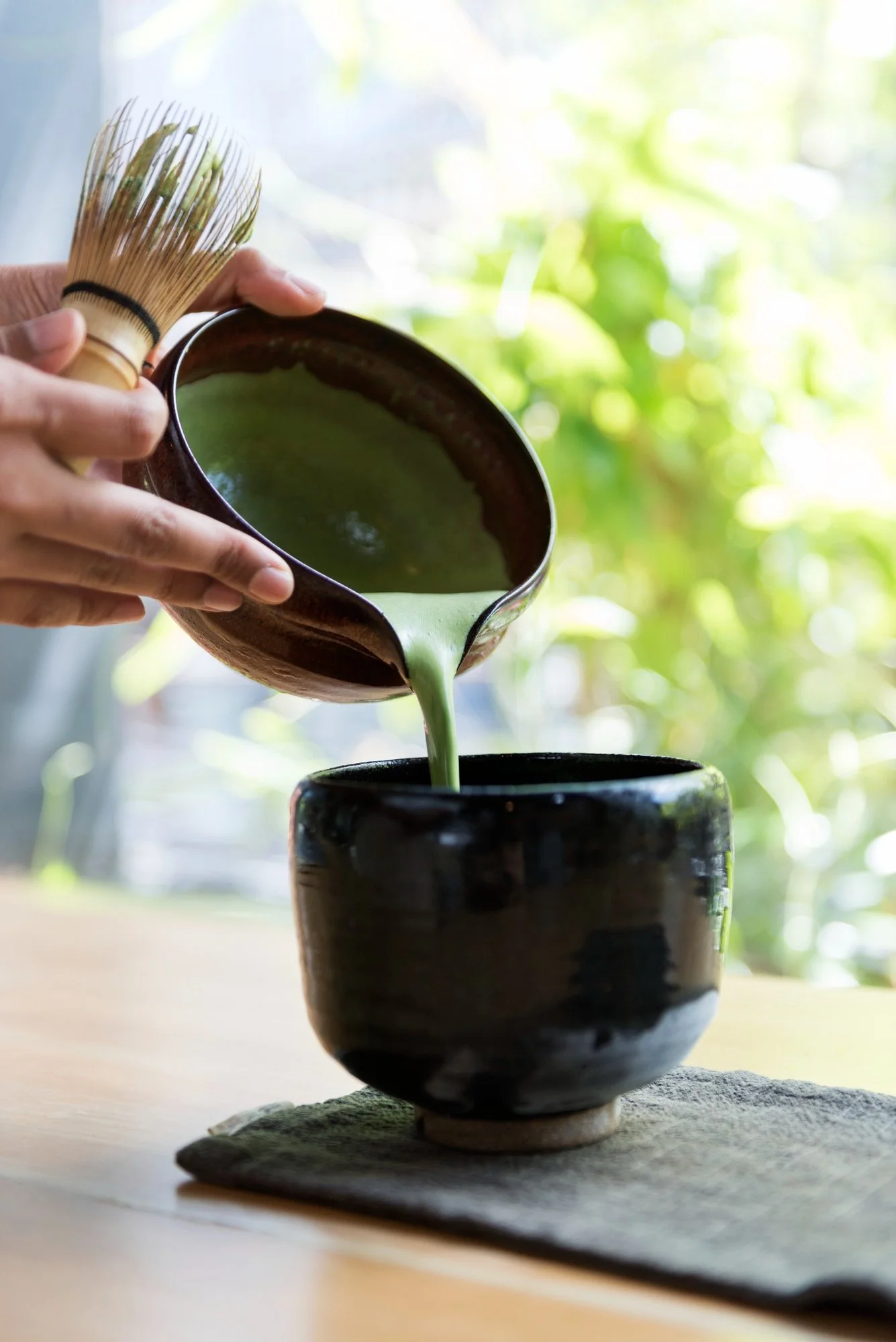A person pouring matcha green tea from a bowl into a black cup, with a bamboo whisk in hand, on a wooden table with a gray cloth. The background shows blurred greenery outside.