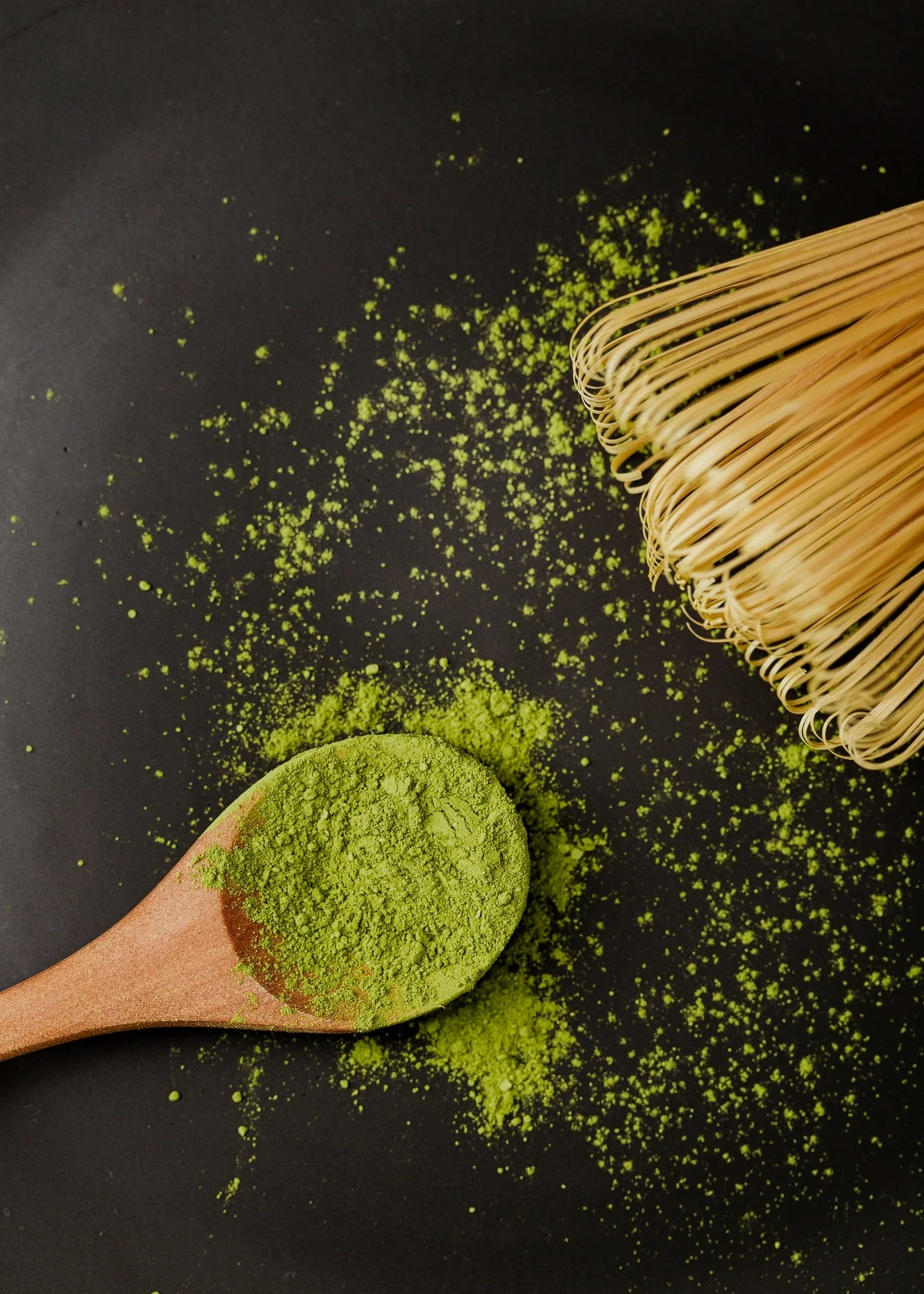 Green matcha powder on a wooden spoon with a bamboo whisk nearby on a dark surface.