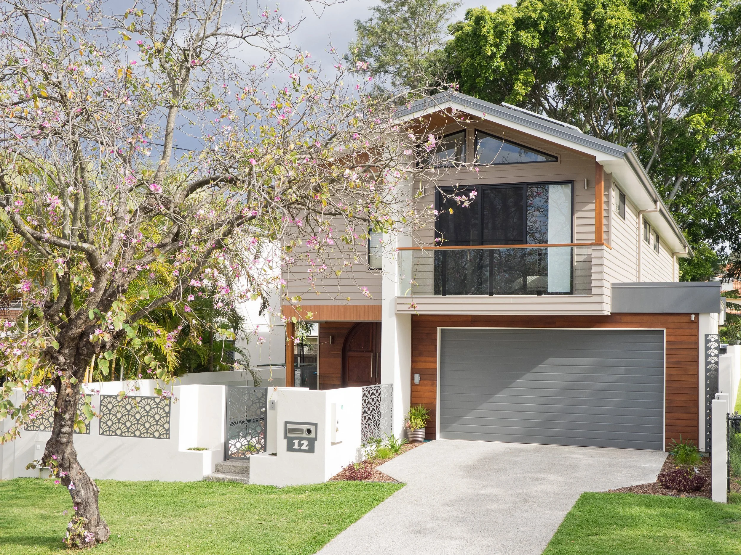 Modern two-story house with gray and brown exterior, large front window, attached garage, and landscaped front yard with flowering tree and green grass.