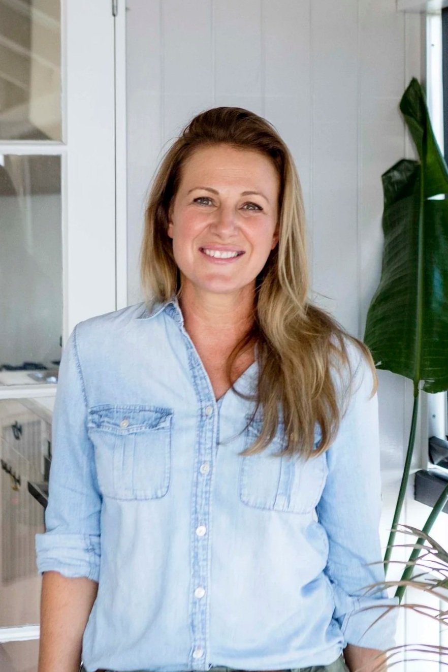A woman with long blonde hair smiling, wearing a light blue denim shirt, standing indoors with a large green plant and white walls in the background.