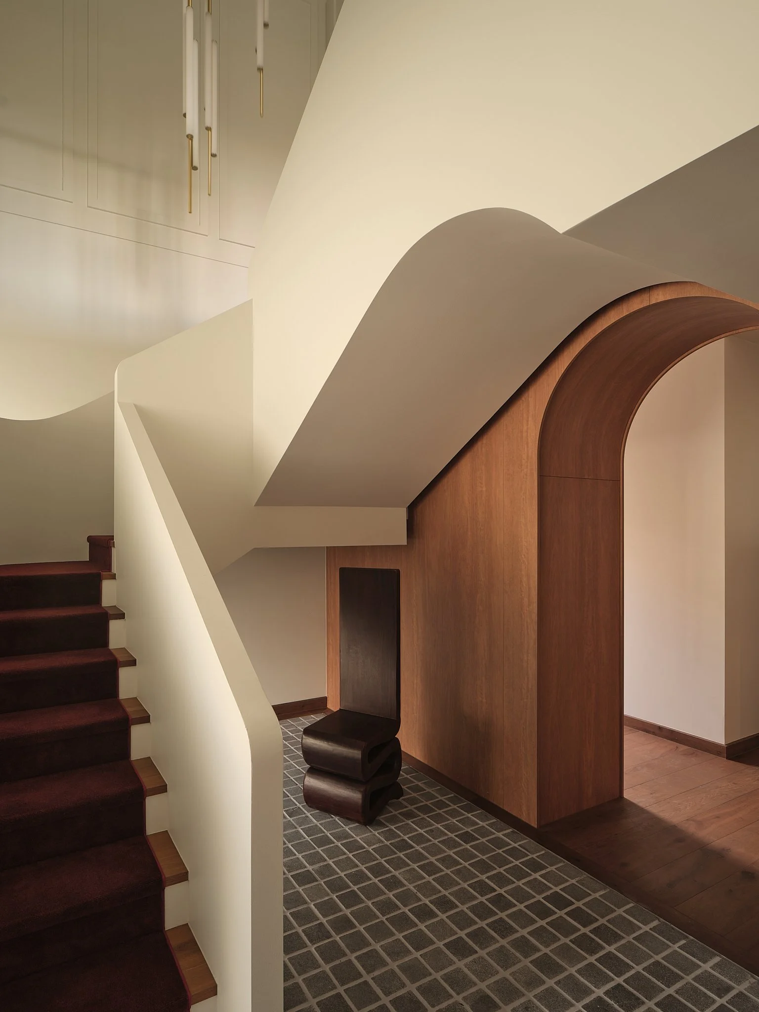 Interior view of a modern staircase with dark carpeted steps, a unique black chair, and wooden structural elements with curved design, modern lighting, and a tiled floor.