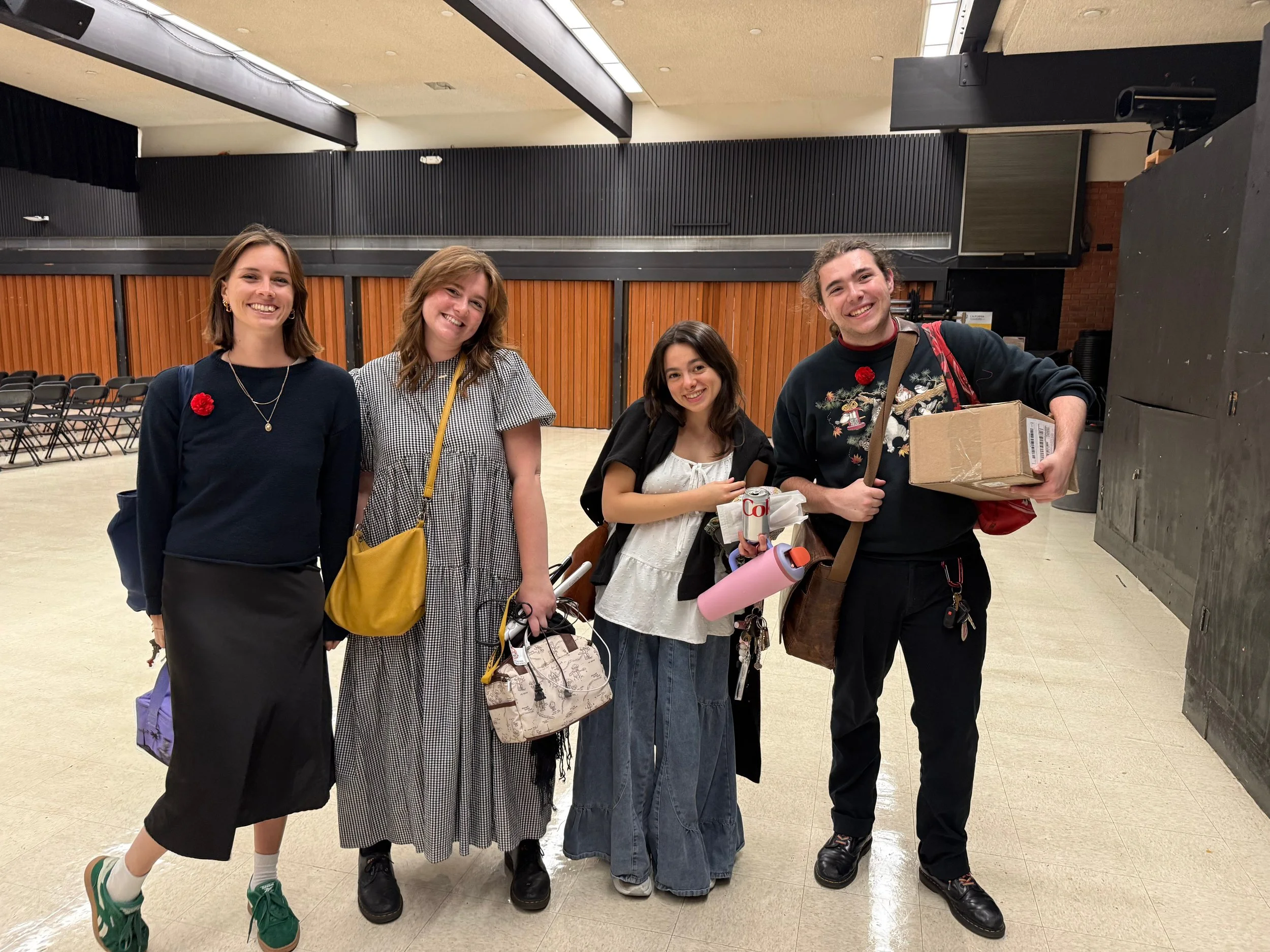 Four young adults standing together in a large room, smiling at the camera, holding various items and bags, with chairs lined up against the wall in the background.
