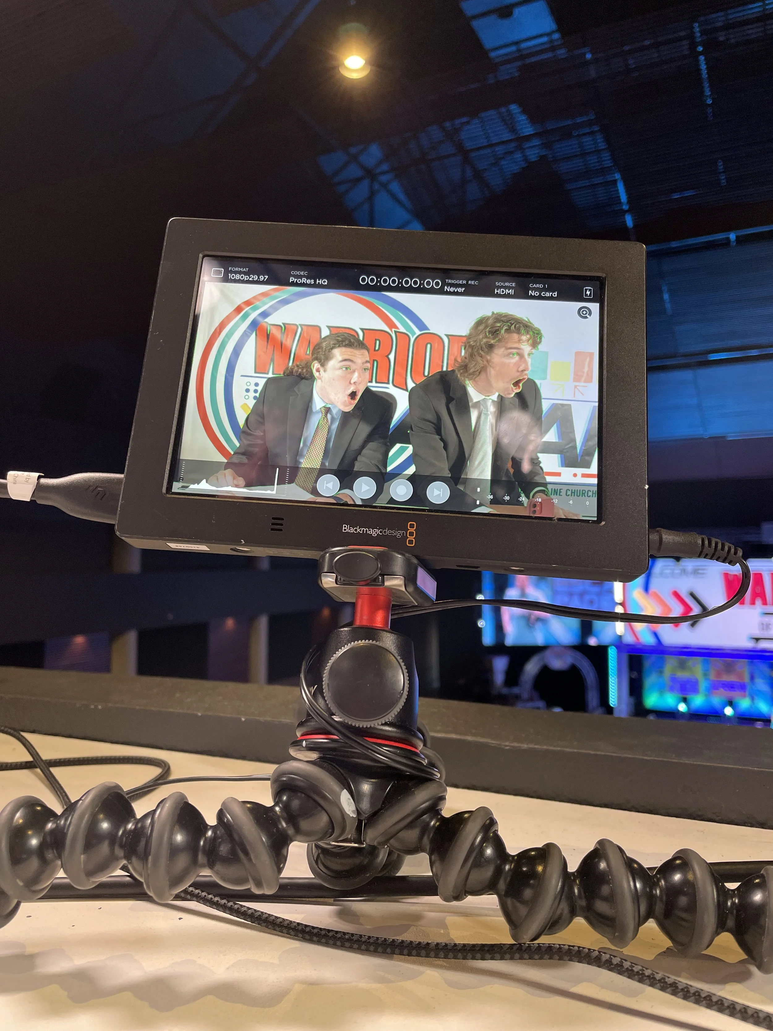 A camera monitor on a tripod captures two men in suits during a performance or presentation, with a colorful background that includes the word "WARRIOR".