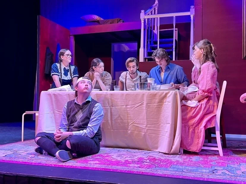 Six people performing a scene on stage in a room with a dining table and a loft bed. One boy is sitting on the floor looking up, while five others sit at the table. The scene is lit with purple and pink stage lighting.