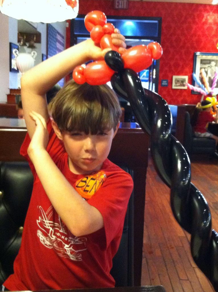 A young boy in a red shirt with a name tag that says 'Ben,' holding a balloon sword with a twisted black and red pattern in a restaurant or party setting.