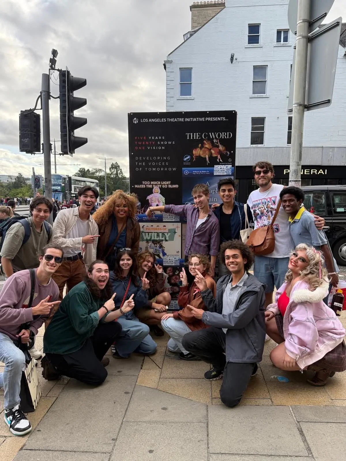 A group of diverse young people smiling and posing in front of a theater poster on a city street, with traffic lights and buildings in the background.