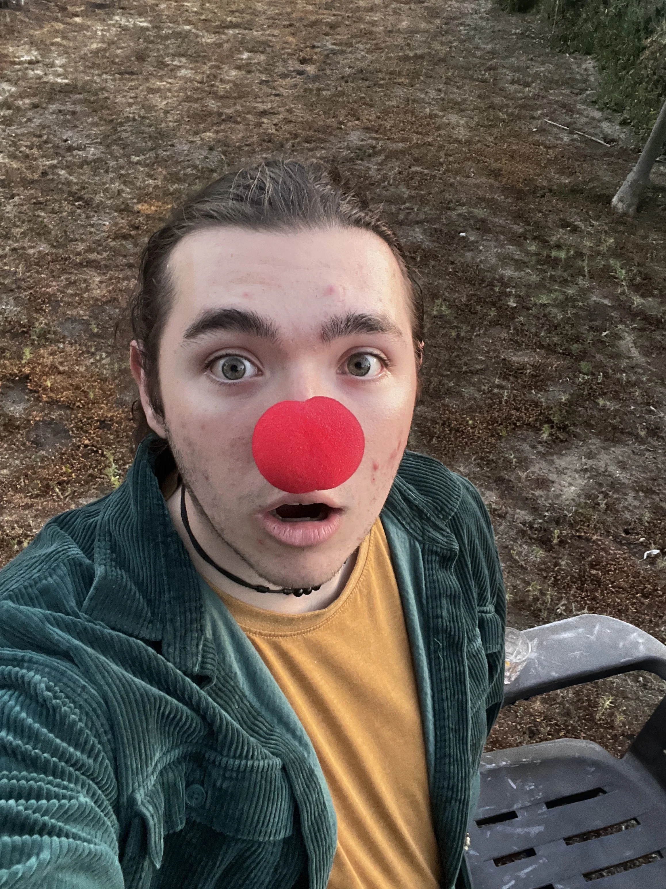 A young person with light skin, dark hair, and a surprised expression wearing a red clown nose, a yellow shirt, and a green jacket, sitting on a park bench.