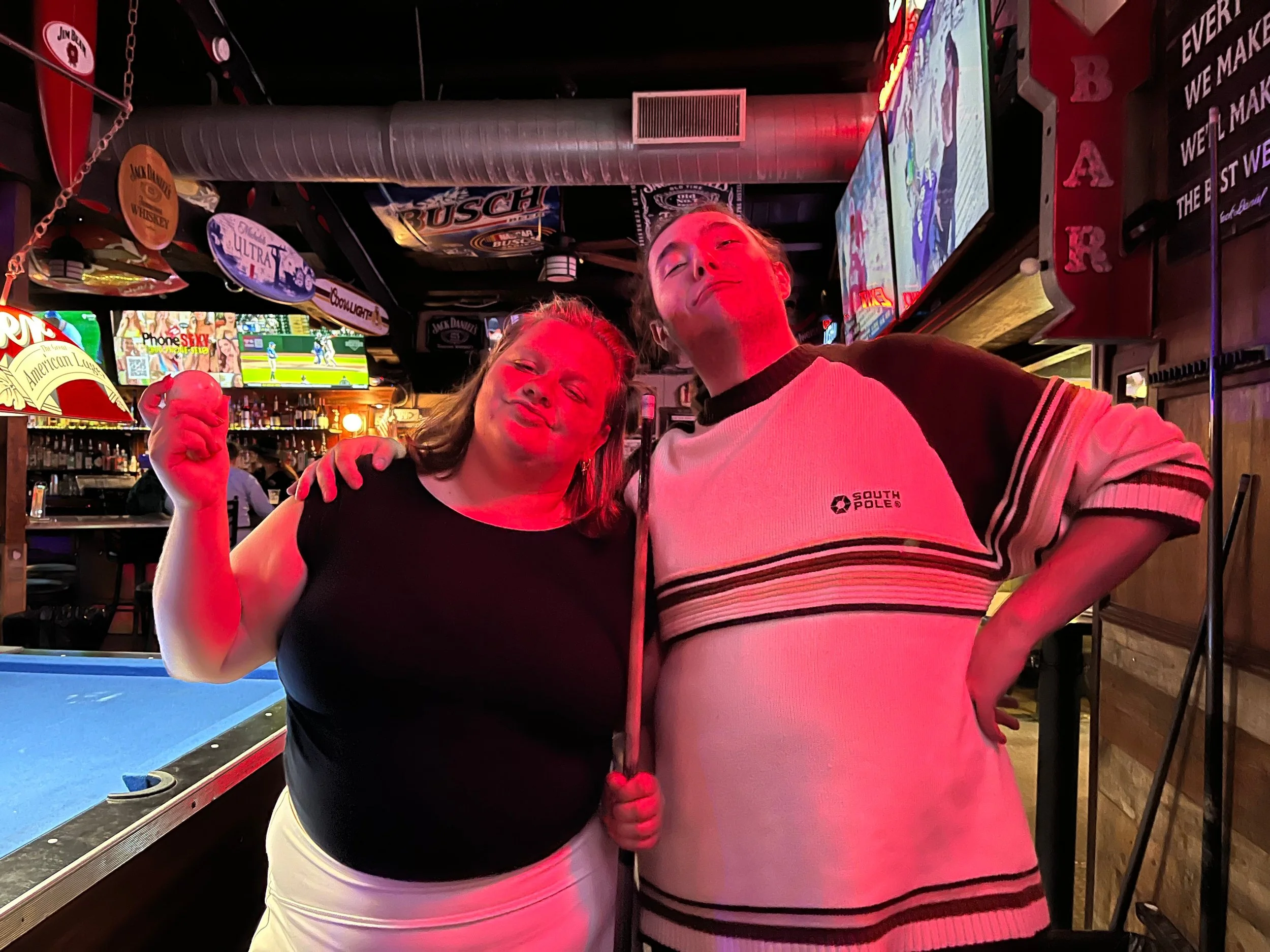 Two friends posing and smiling with relaxed expressions inside a bar, with a pool table and neon signs in the background.