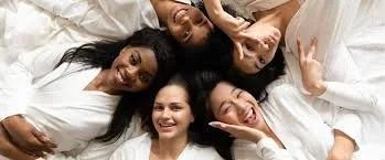 Five women lying on a bed, smiling, with their heads close together.