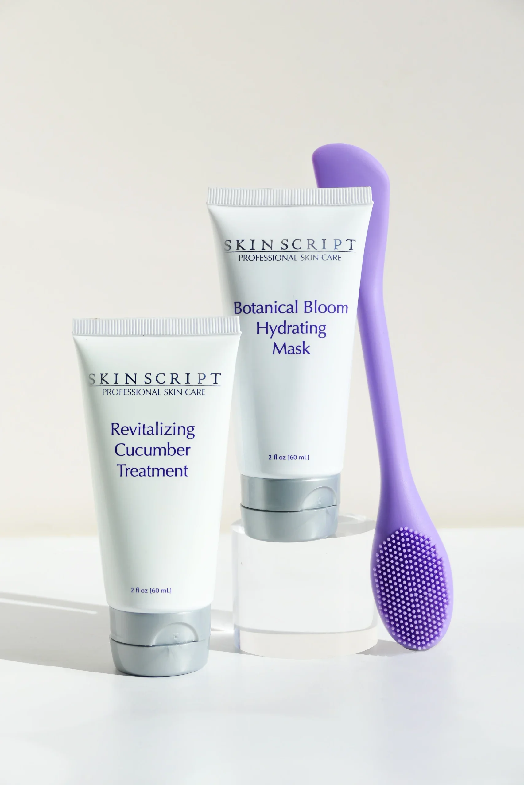 Two skincare treatment tubes labeled 'Revitalizing Cucumber Treatment' and 'Botanical Bloom Hydrating Mask' with a purple silicone face brush on a clear stand, set against a plain background.