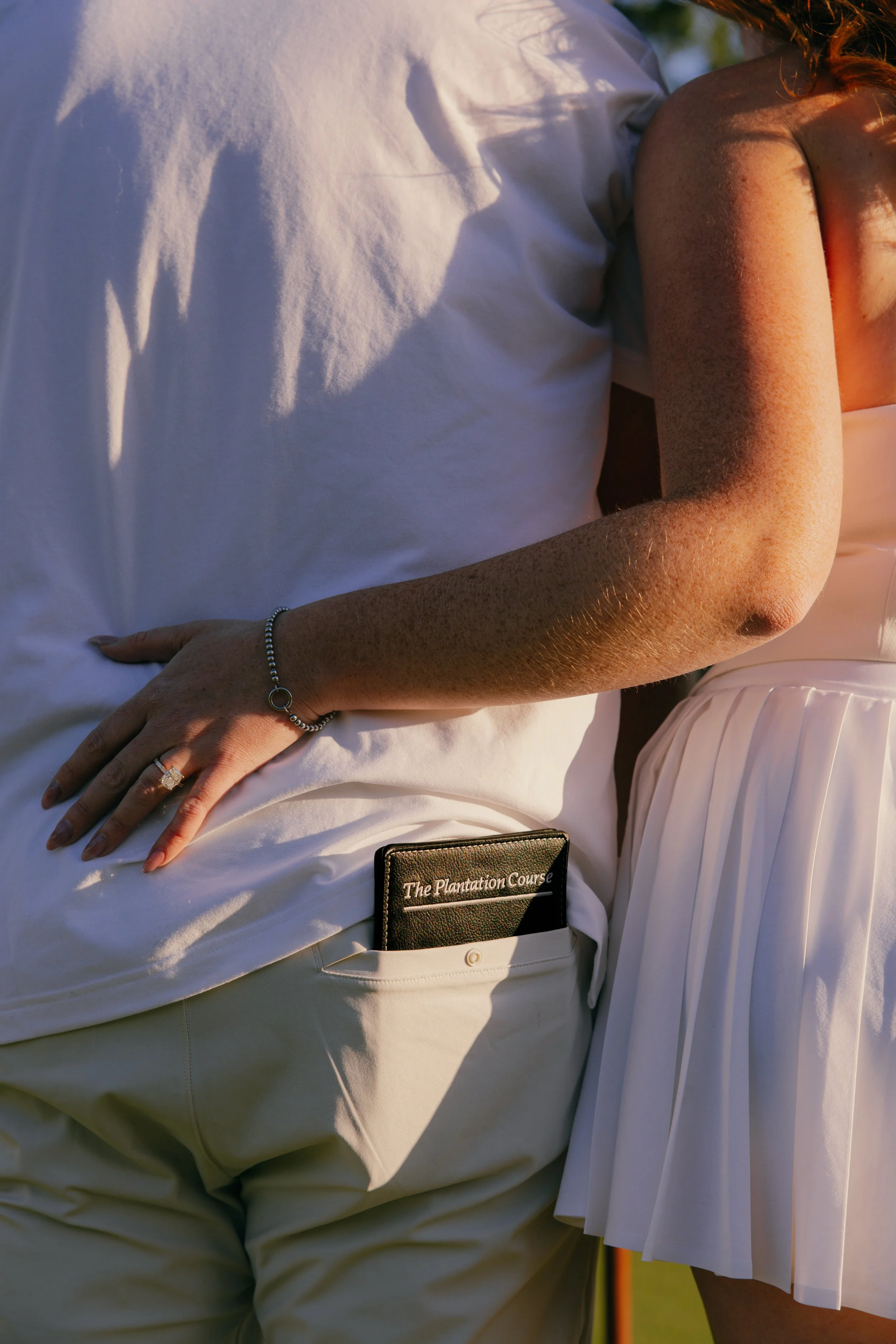 Close-up photo of a person in a white shirt and beige pants, lying down with their hand resting on their hip. A book titled 'The Plantation Course' is visible in the back pocket of the pants. The background shows part of a woman wearing a pleated white skirt.