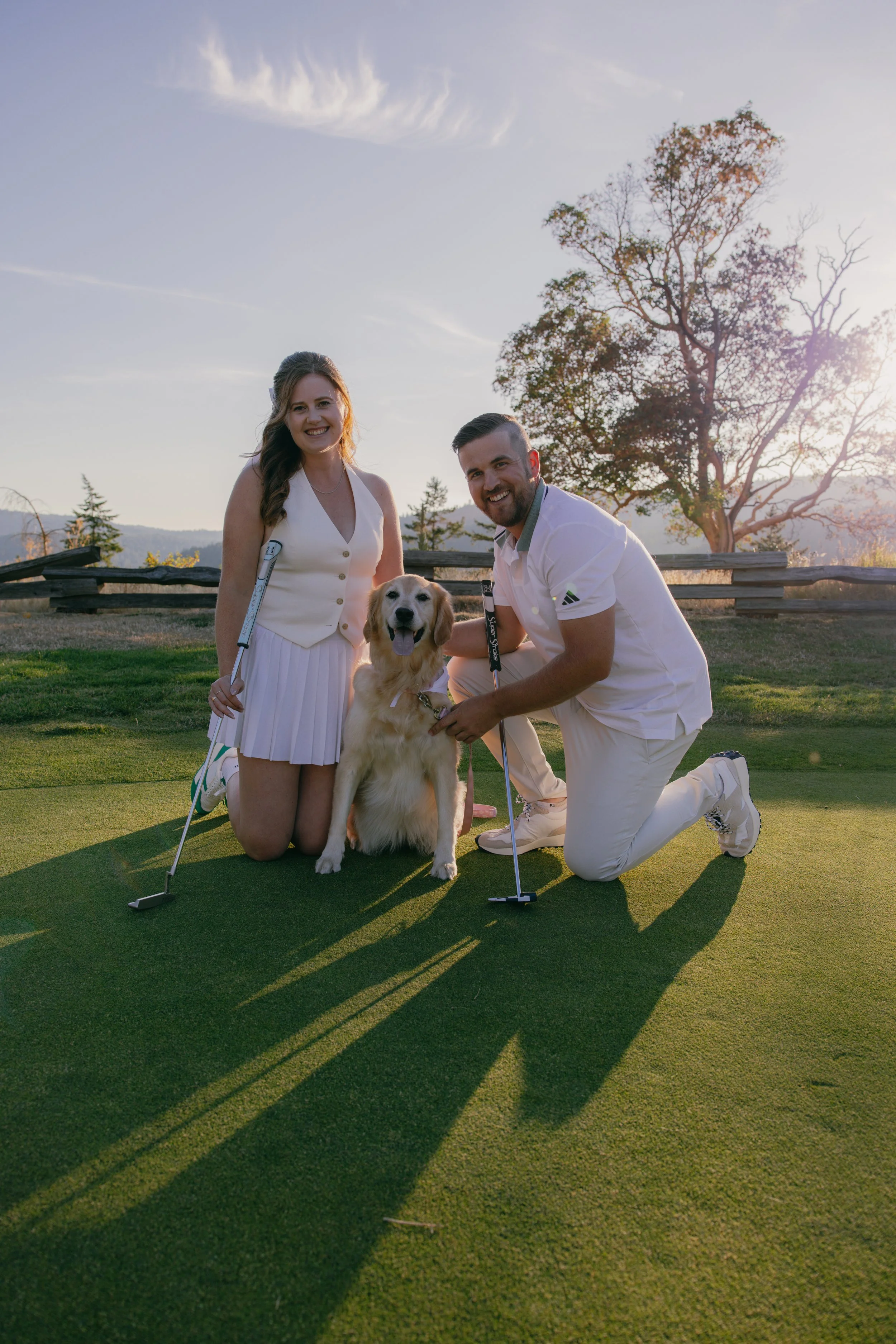 A couple kneeling on a golf course with their dog, holding golf clubs, during sunset.