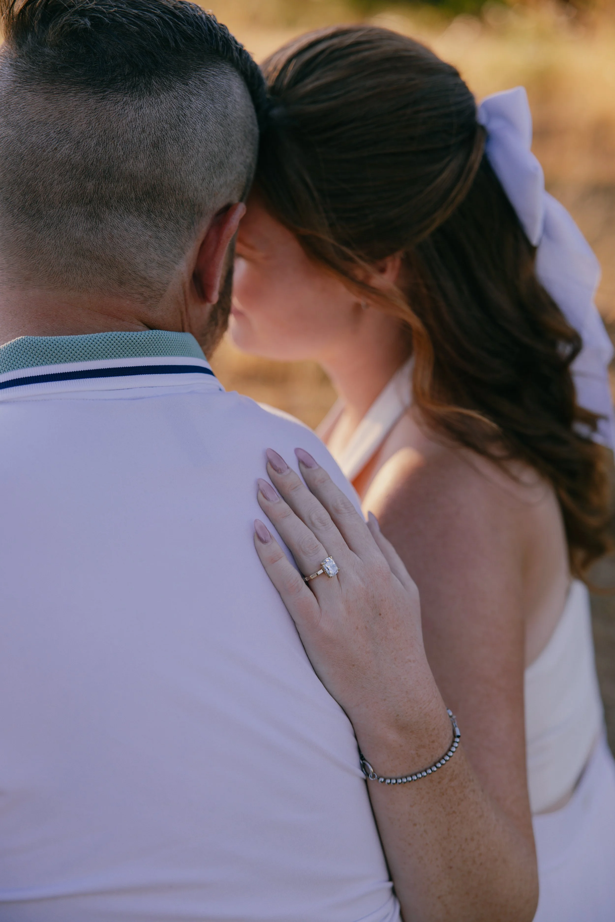A couple sharing a kiss outdoors, with the woman's left hand on the man's shoulder displaying a diamond engagement ring.
