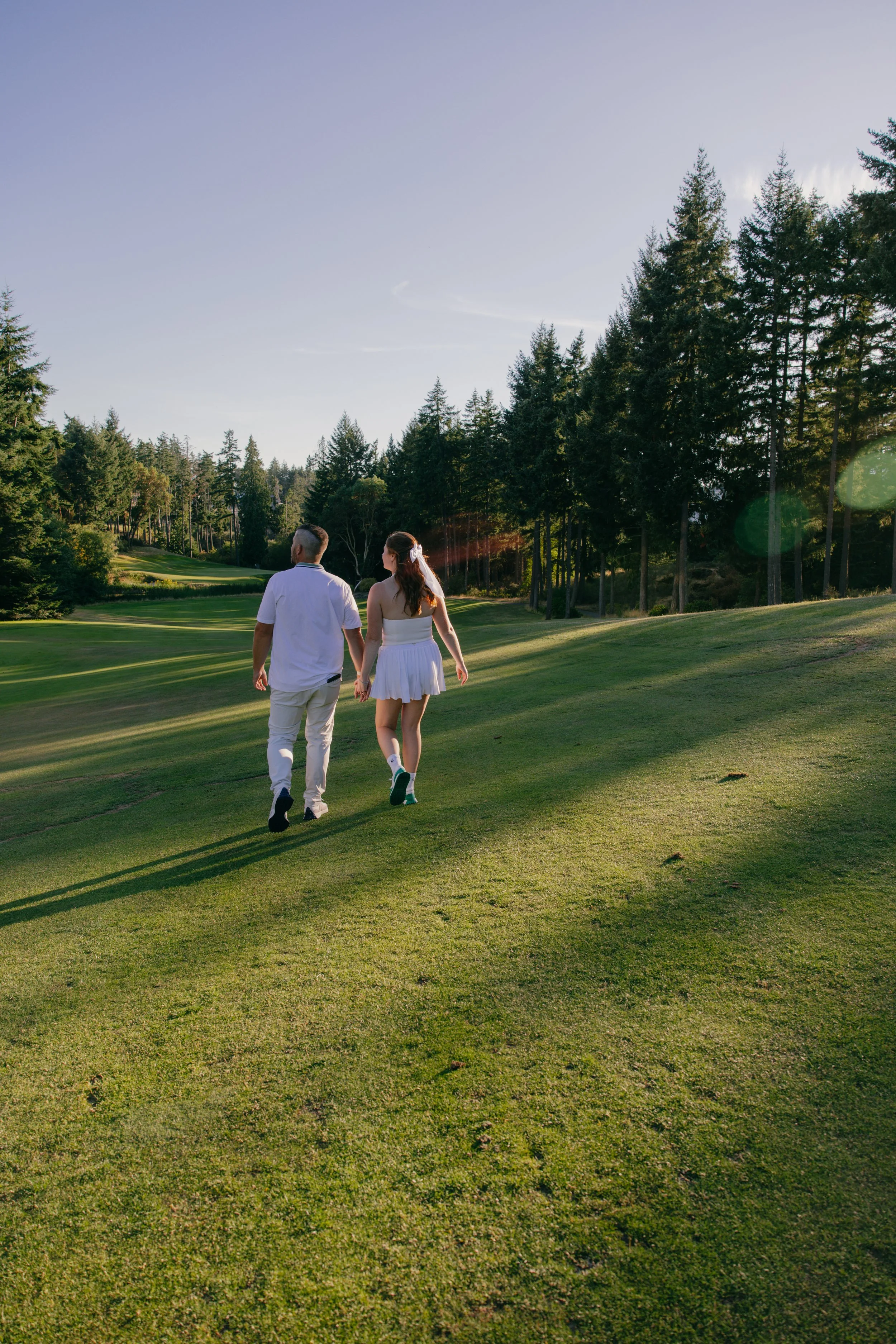 A couple walking hand in hand on a golf course with tall trees in the background during late afternoon sunlight.