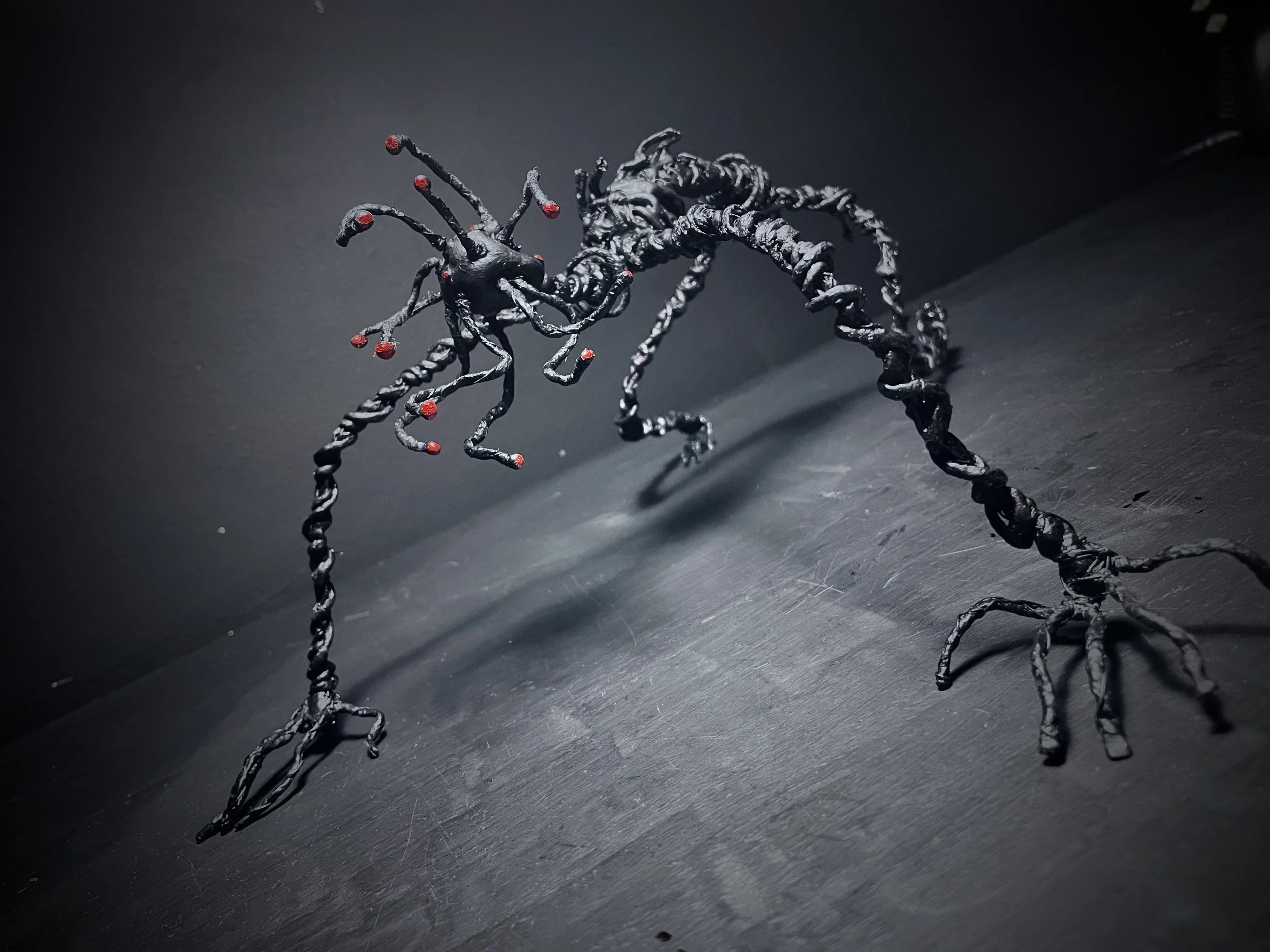 Artistic sculpture resembling a crawling creature with a twisted, black, metallic body and red-tipped antennae on a dark surface.