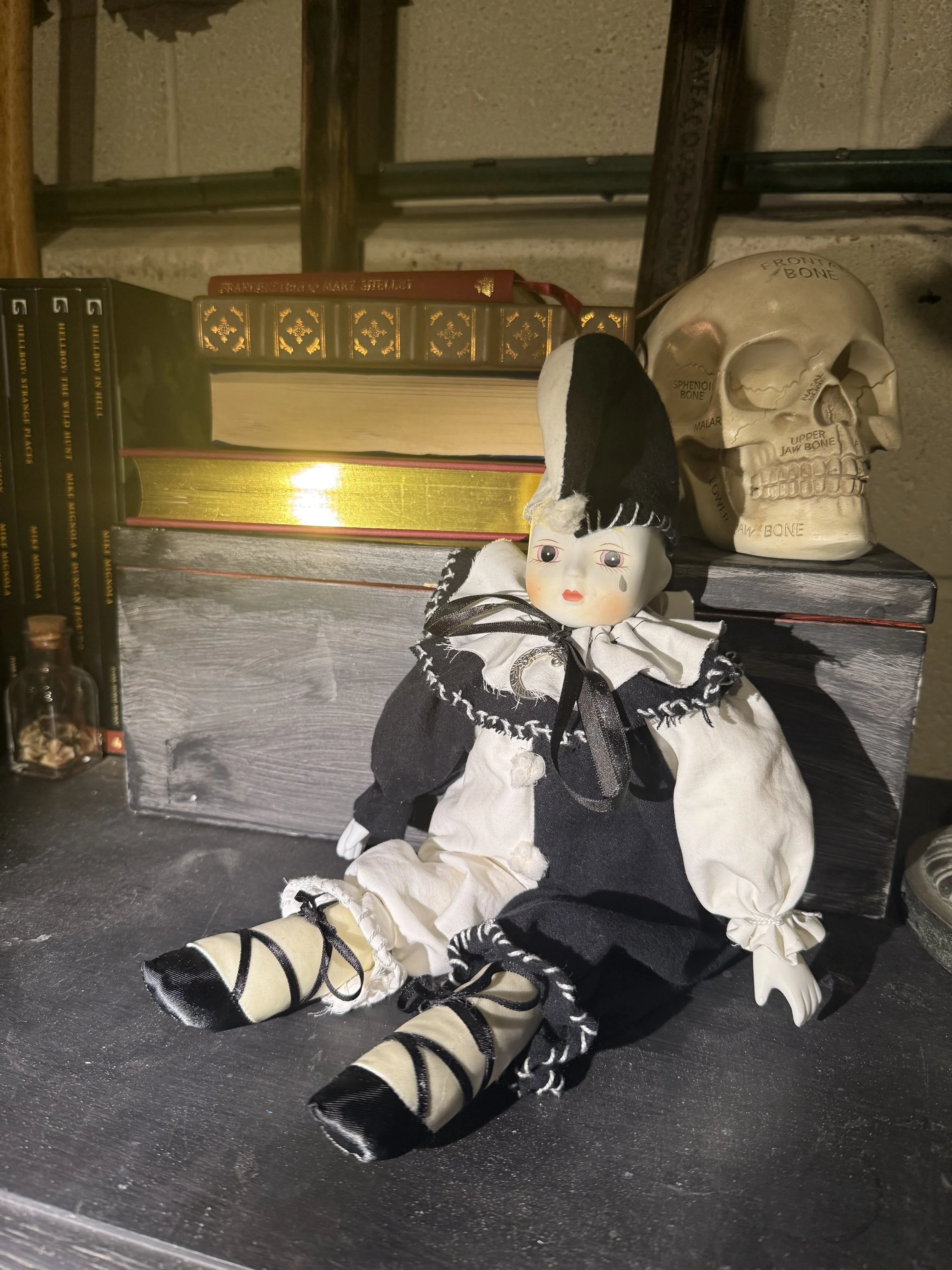 A vintage doll dressed in black and white with a tear drop painted on cheek, sitting on a dark surface next to a skull model, books, and a small jar.