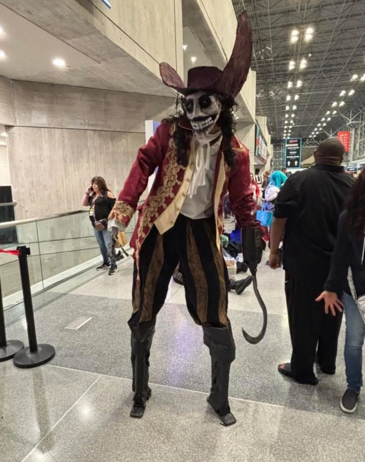A person dressed as a skeleton pirate with a large hat, red coat, striped pants, and a tail, standing in an indoor public space, possibly a convention or airport, with other people in the background.