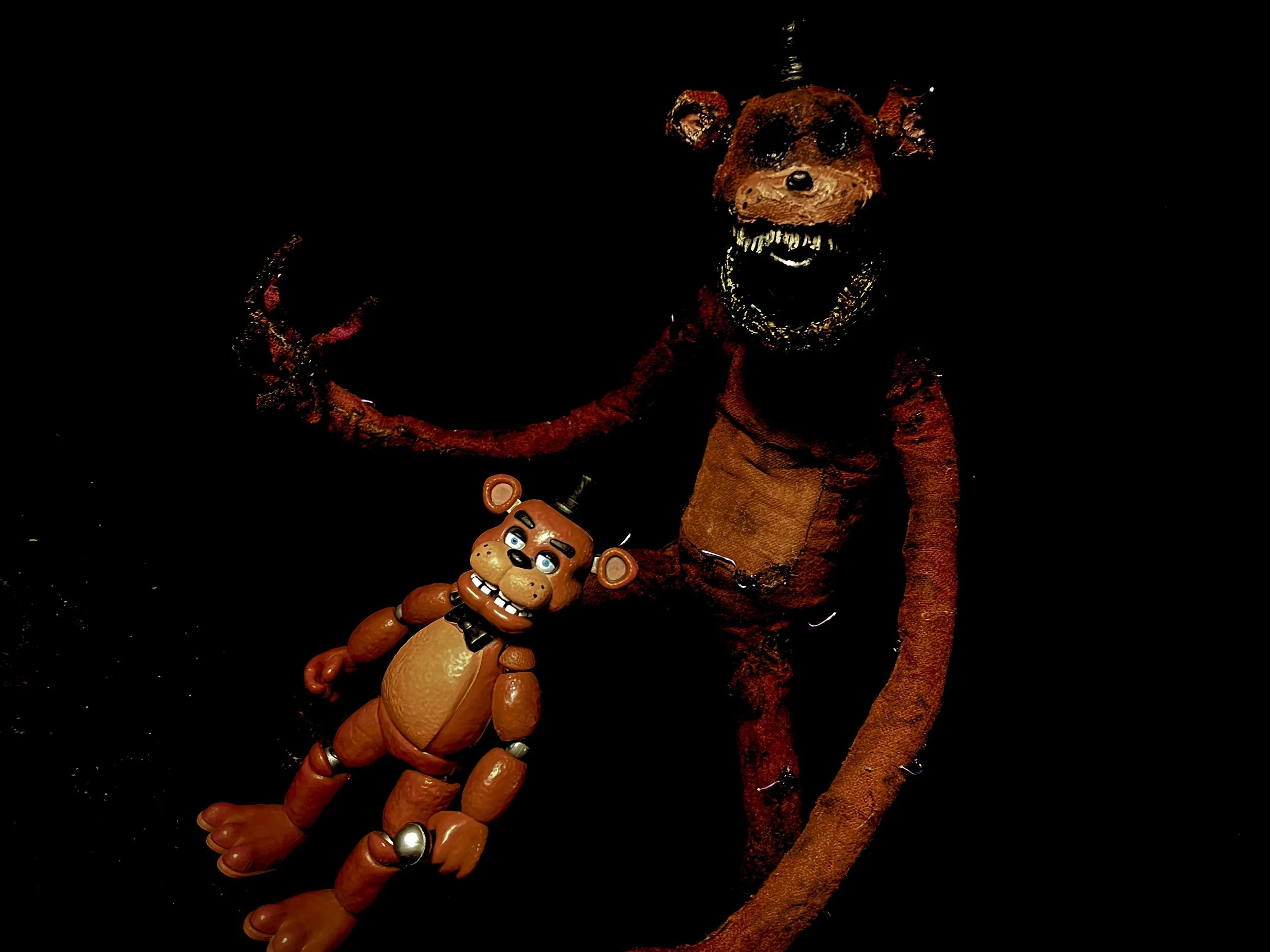A plush toy of a red or orange fox with a chainsaw in its paw, and a life-size puppet of a humanoid fox with a wide grin, sharp teeth, and dark eyes, both set against a black background.