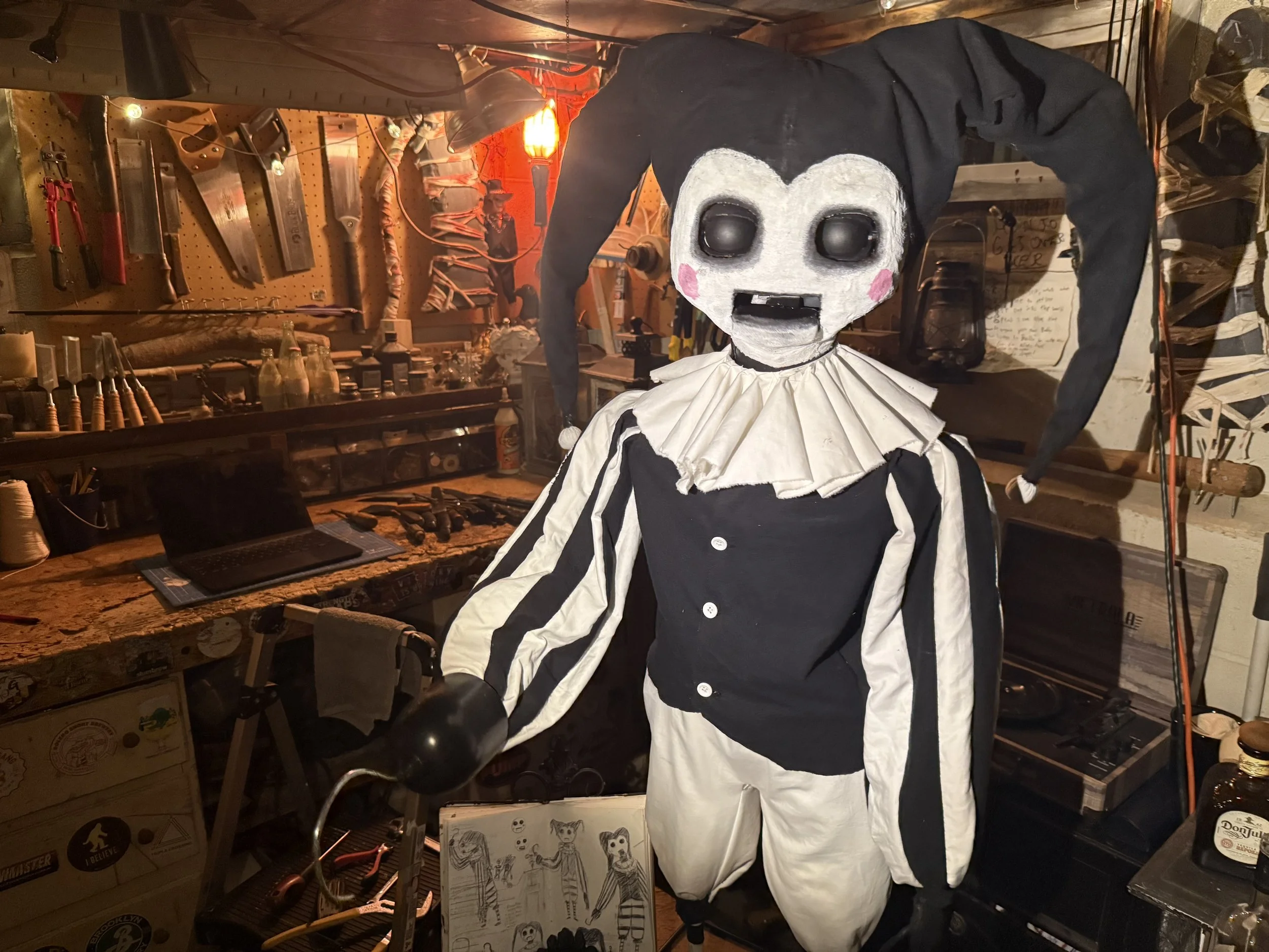 A spooky clown puppet with a white face, pink cheeks, and black eyes, dressed in a black and white costume with a ruffled collar, standing in a cluttered workshop with tools, bottles, and drawings in the background.