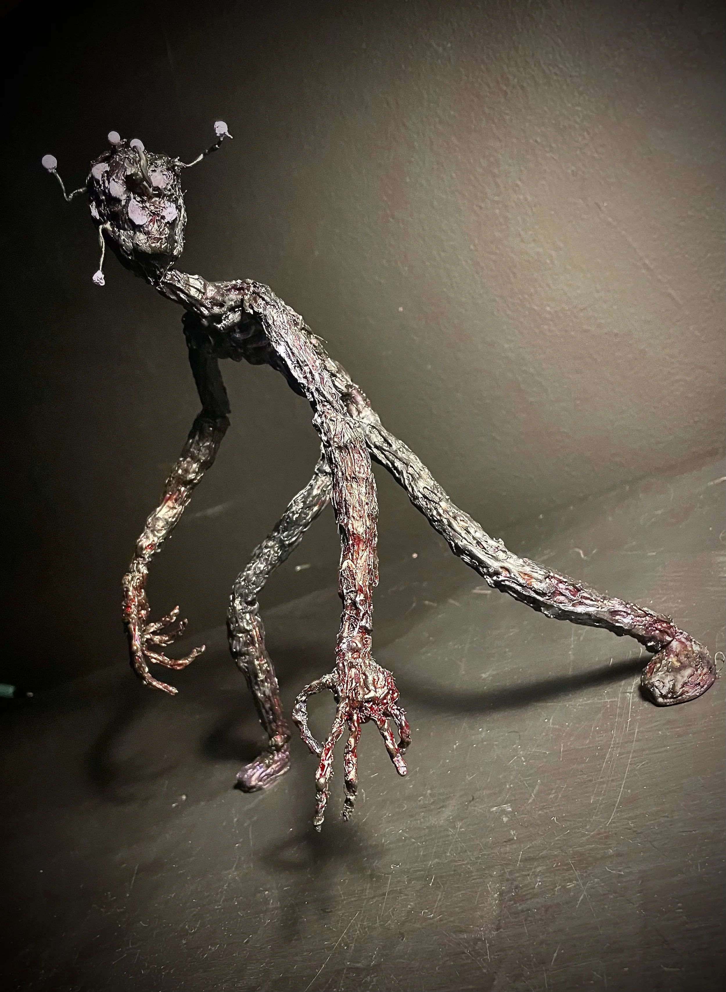 A twisted, metallic sculpture resembling a humanoid figure with long limbs, a textured surface, and antenna-like protrusions on its head, placed on a dark surface against a gray wall.